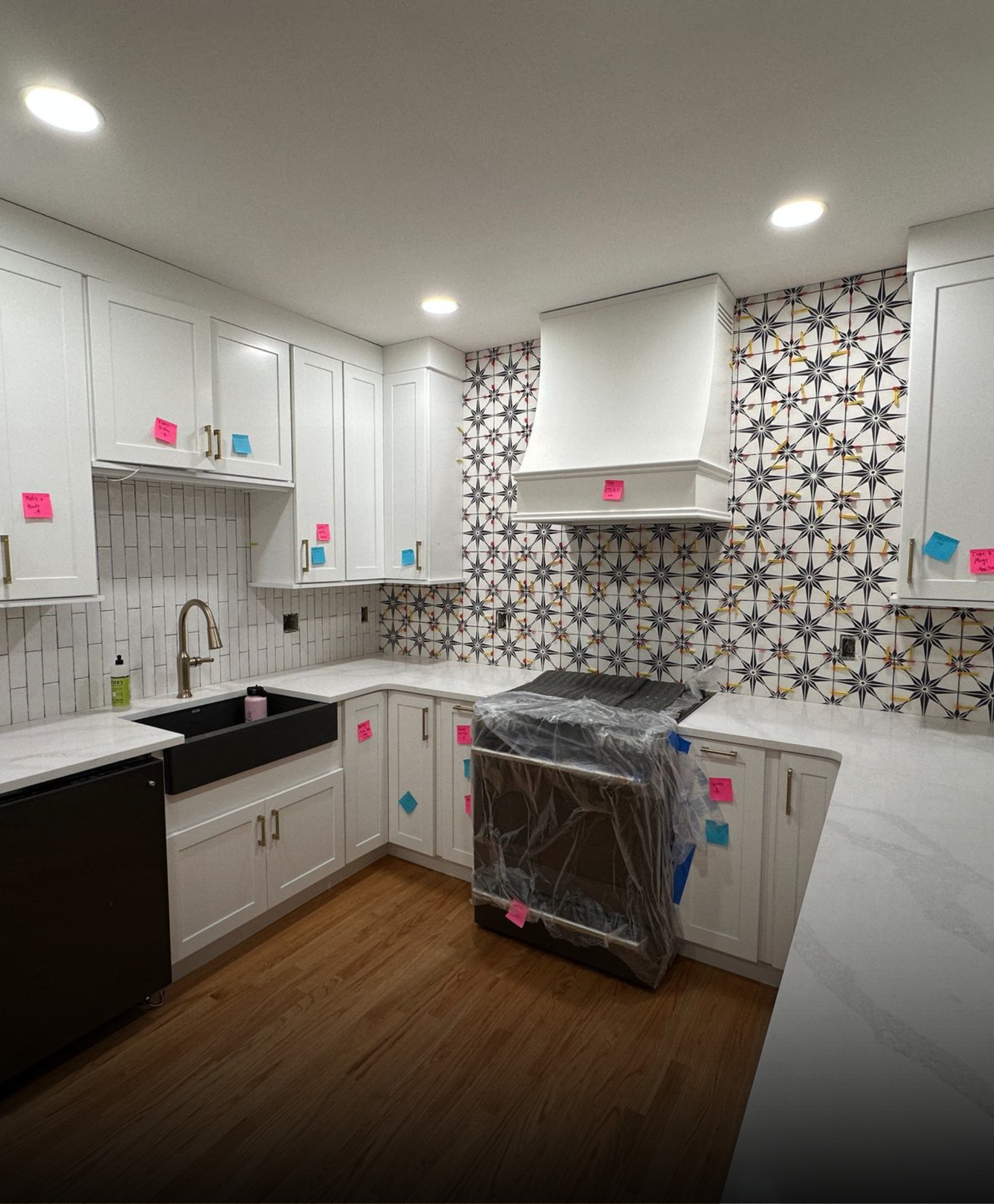 A kitchen with white cabinets, a black sink, a patterned tile backsplash, a stove wrapped in plastic, and wood flooring.