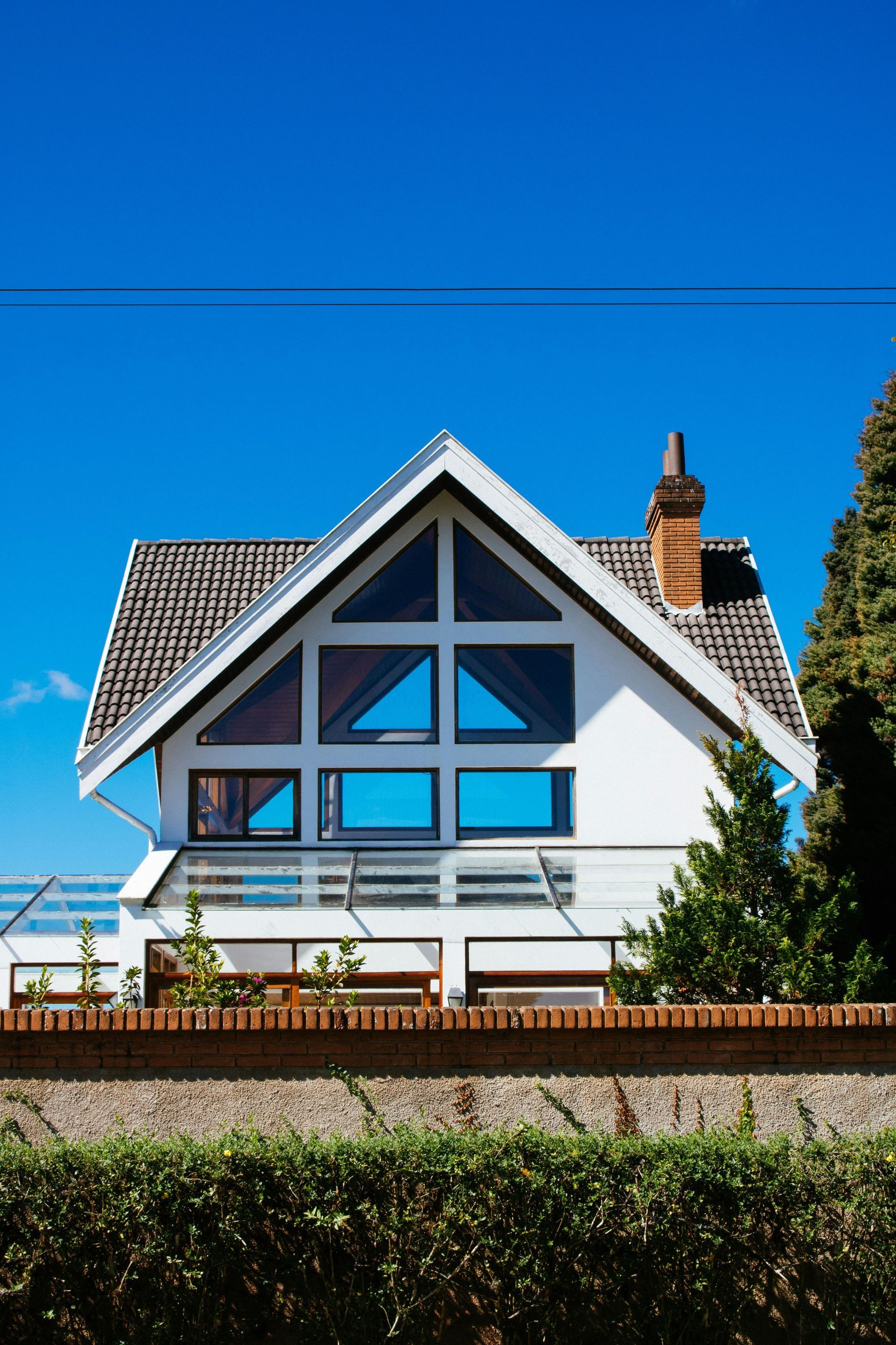 White house with triangular window, blue sky. Brick fence and green hedge in front.