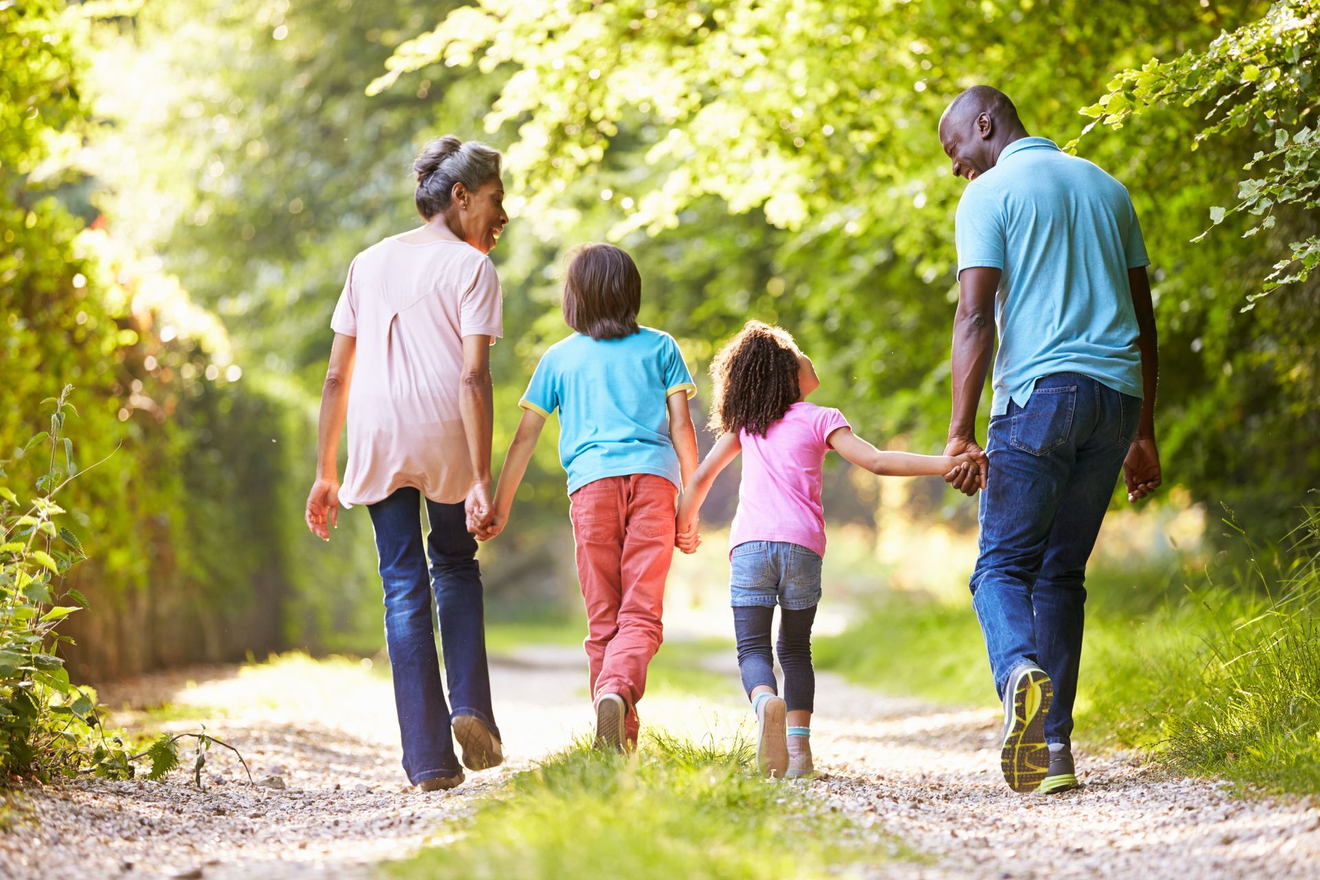 A family of four walking hand-in-hand down a sunlit, tree-lined path in the woods.