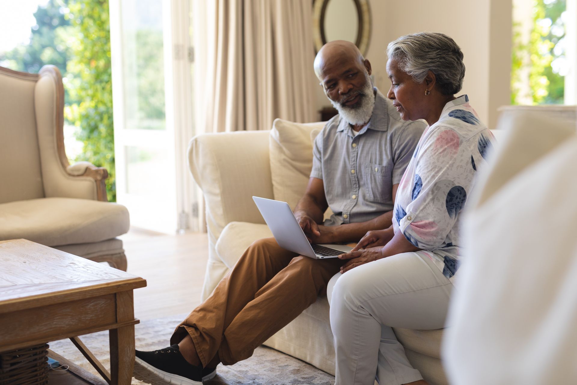 An older couple sits together on a couch in a bright living room, looking at a laptop computer held on their laps.