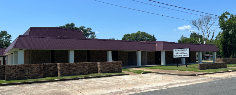 A one-story building with a maroon roof and stone exterior, featuring a white sign and white columns under a long overhang.