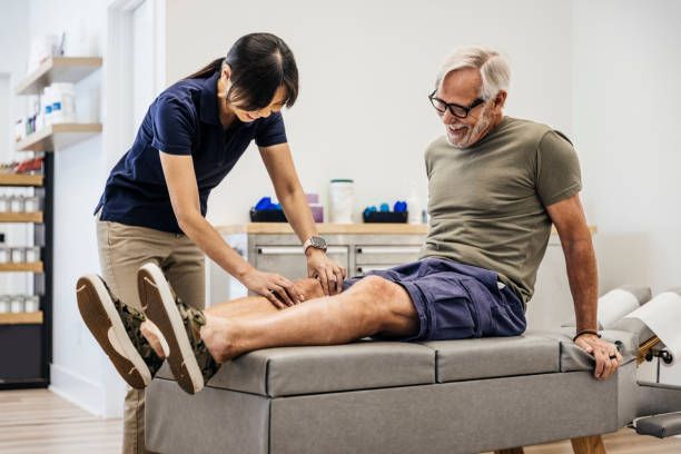 A physical therapist examining a senior man's knee on a bench. She wears a blue shirt, he a green one, in a light-filled room.