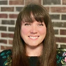 A woman with long hair and bangs is smiling in front of a brick wall.