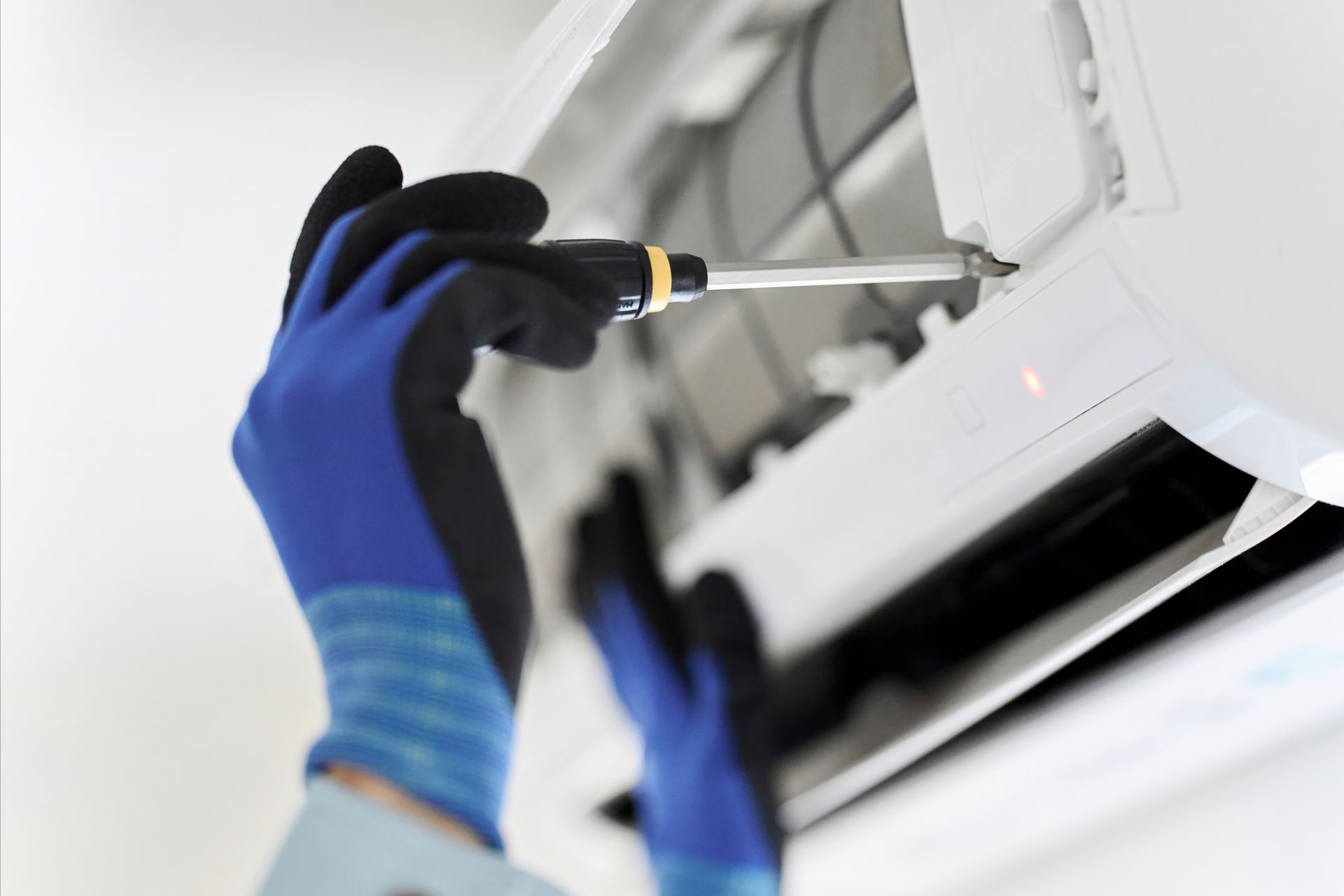 A person wearing blue and black work gloves uses a screwdriver to repair a wall-mounted air conditioner.