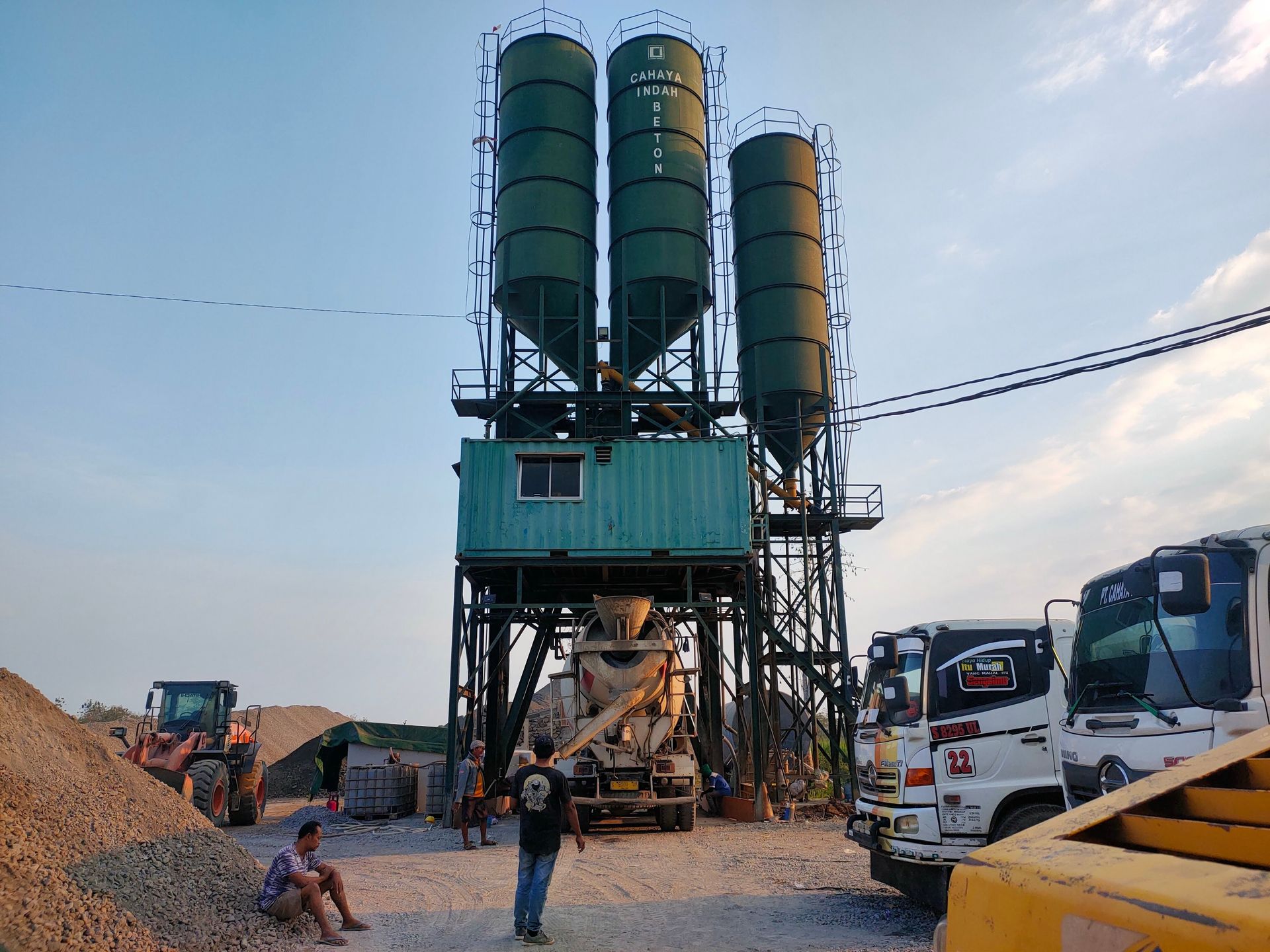 Concrete batch plant with three silos, a concrete mixer, trucks, and workers on site.