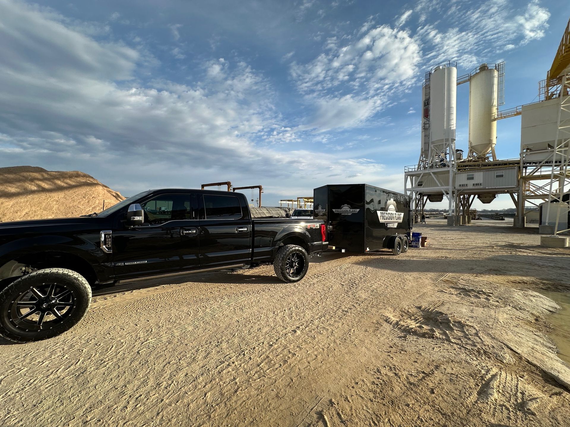 Black pickup truck towing a trailer on a gravel lot near industrial silos under a cloudy sky