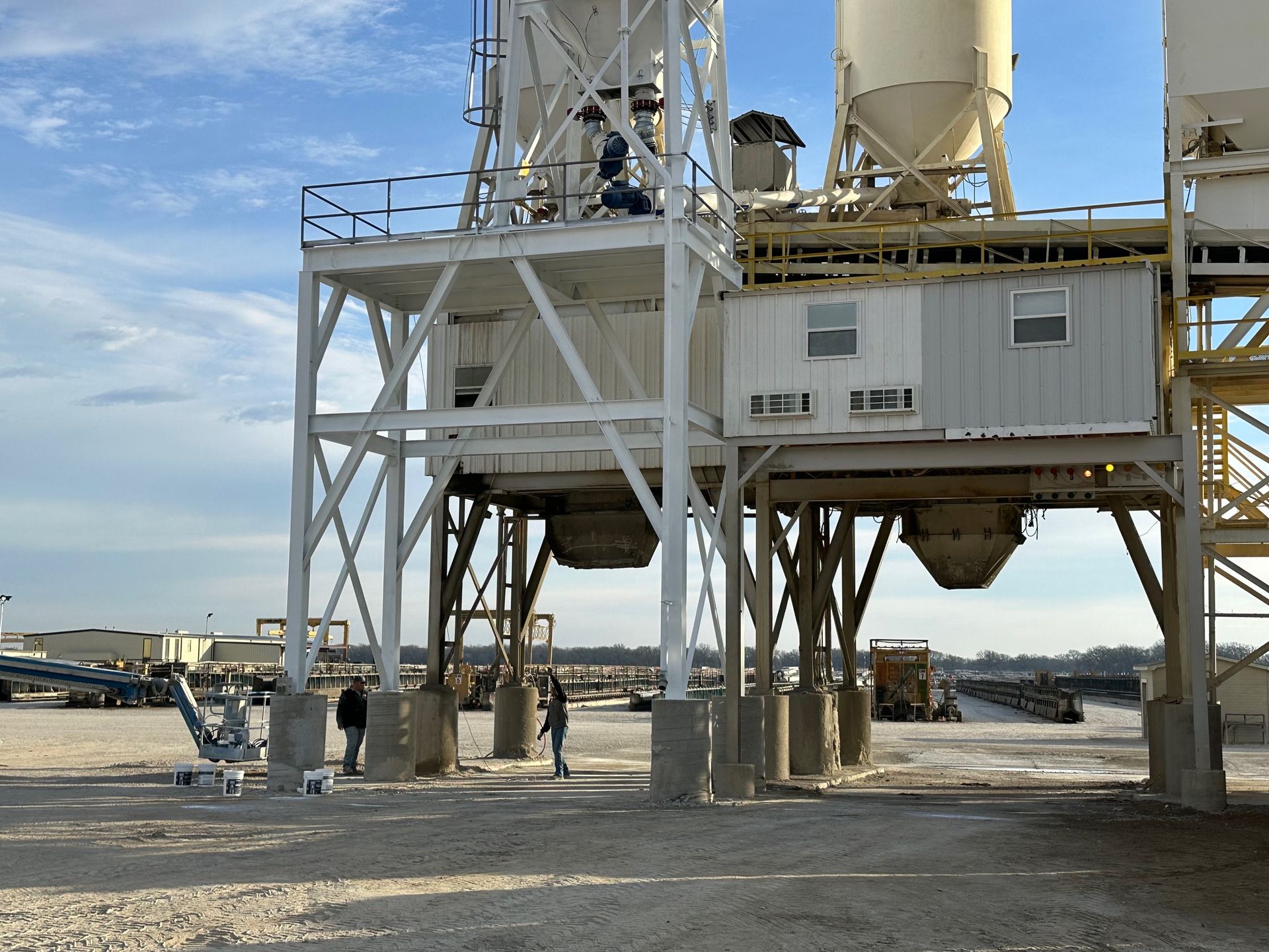 Concrete plant with silos, metal structure, and workers standing in a gravel lot