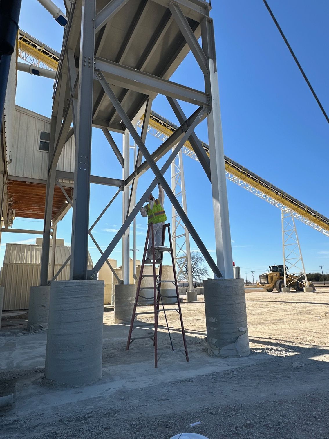 Person on a ladder painting a steel support structure at a construction site; blue sky in background