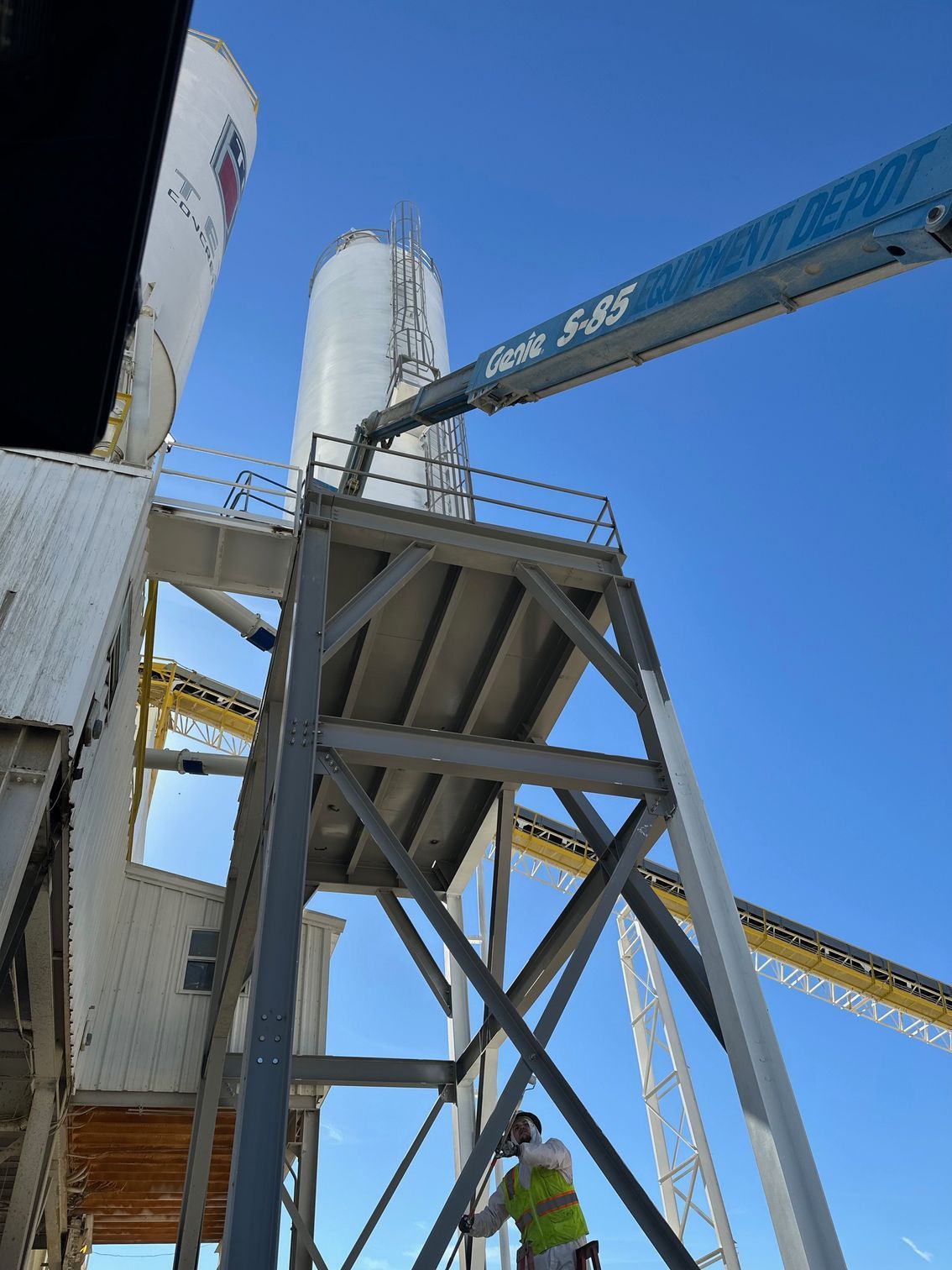 Construction worker near elevated platform, silo, and blue Genie lift against a bright sky