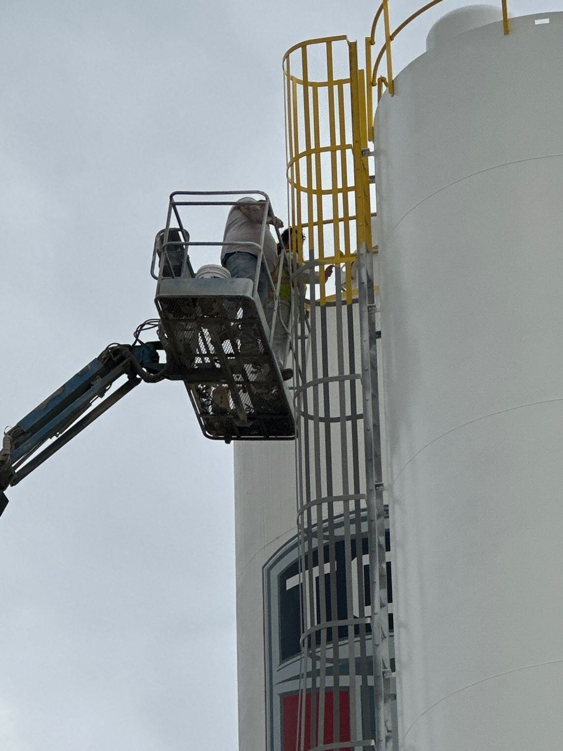 A worker in a lift basket is near a tall white cylindrical structure with a ladder and safety cage