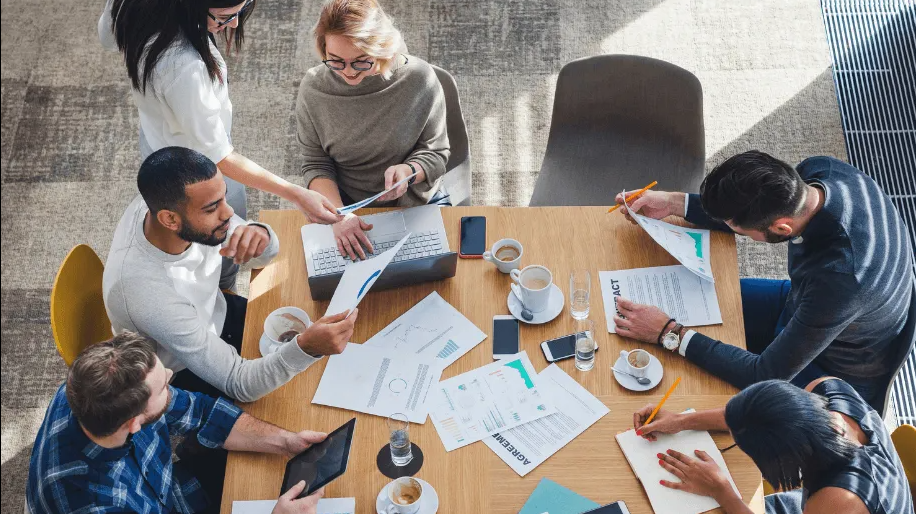 Two computer monitors on a wooden desk with a keyboard, phone, and plant. The left screen shows design layouts.