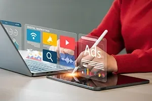 Person in red shirt using a laptop and tablet, digital marketing icons overlaid on screen, and stylus in hand.