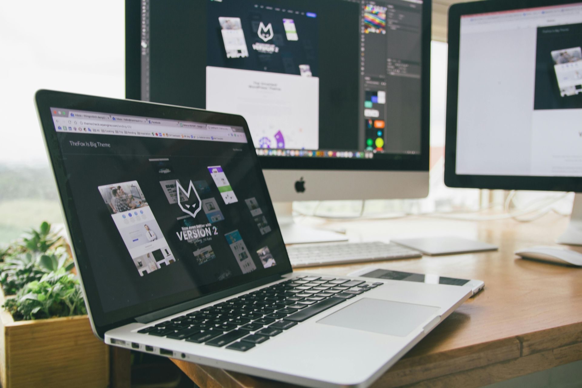 Laptop and two monitors displaying website designs on a wooden desk with a plant.