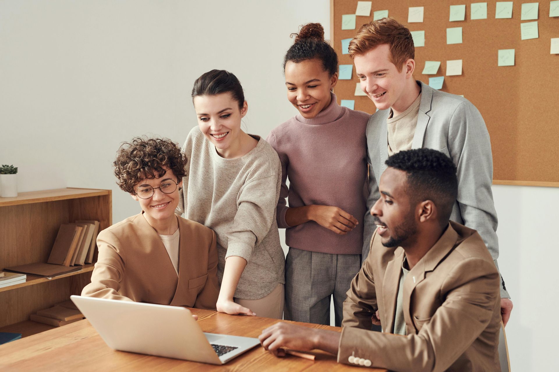 Five coworkers gathered around a laptop, smiling. Light-colored office setting with sticky notes on the wall.