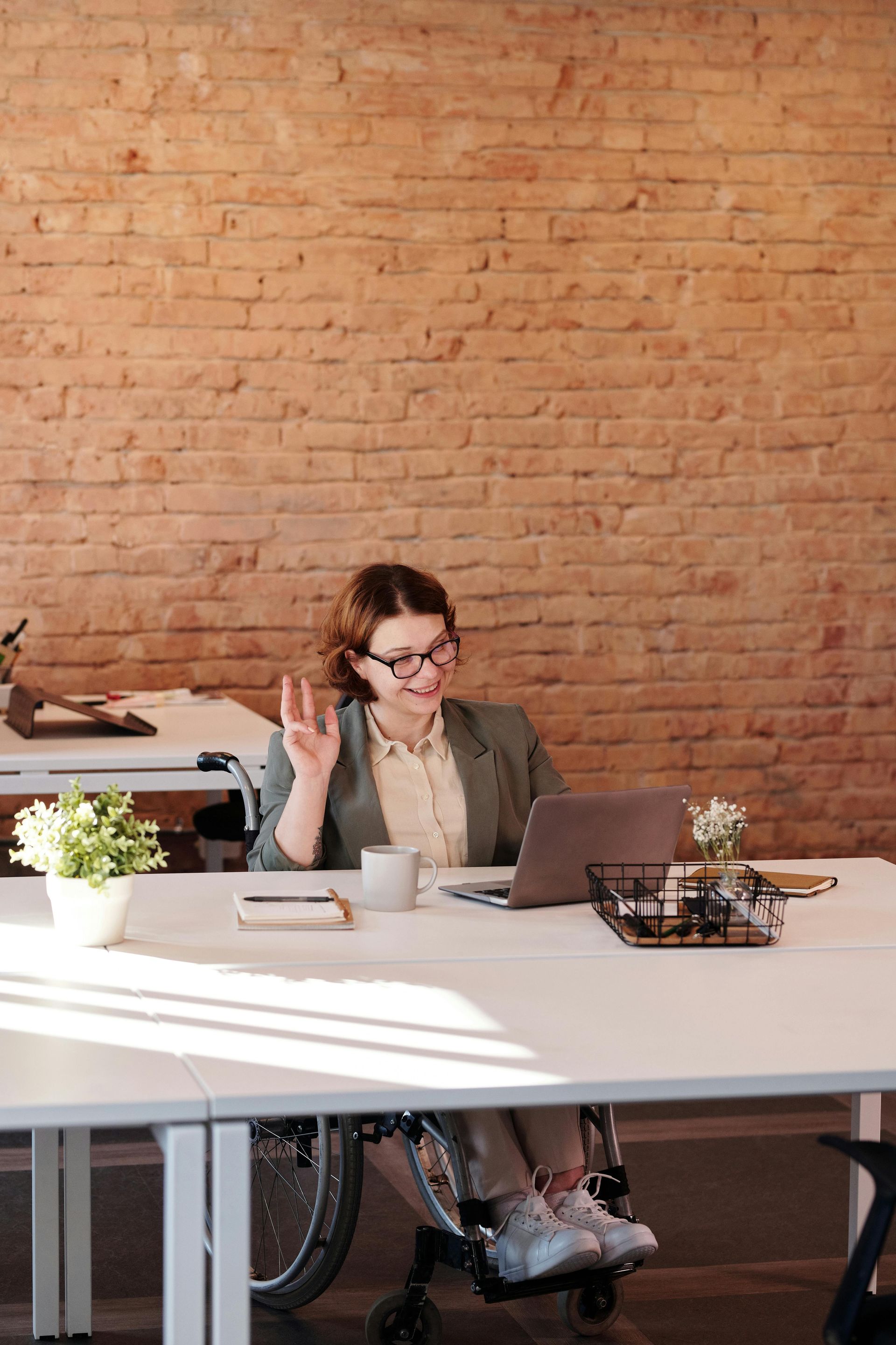 Woman in a wheelchair waves during a video call in an office with brick wall.