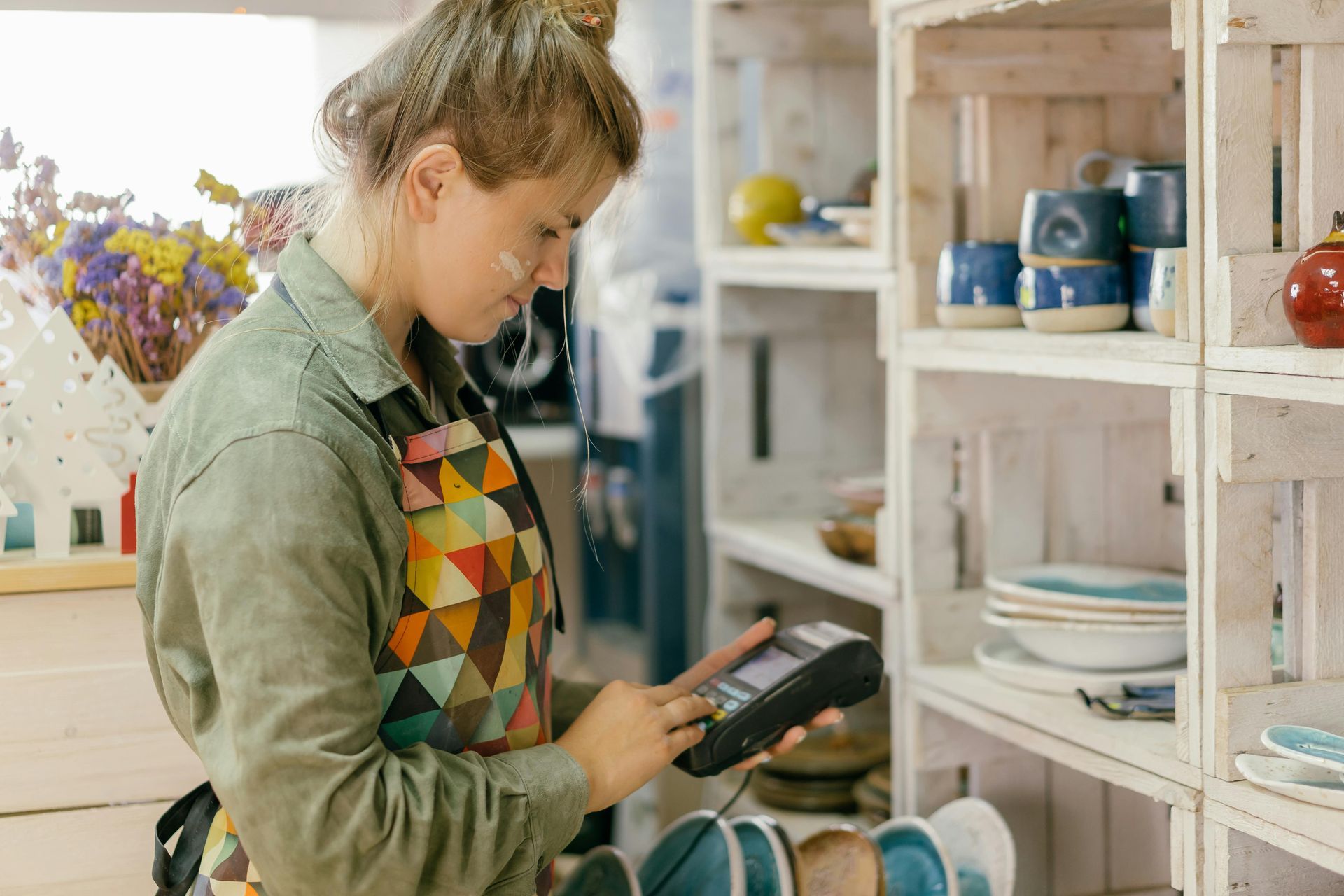 Woman in apron using a payment device in a shop with shelves of ceramic goods.