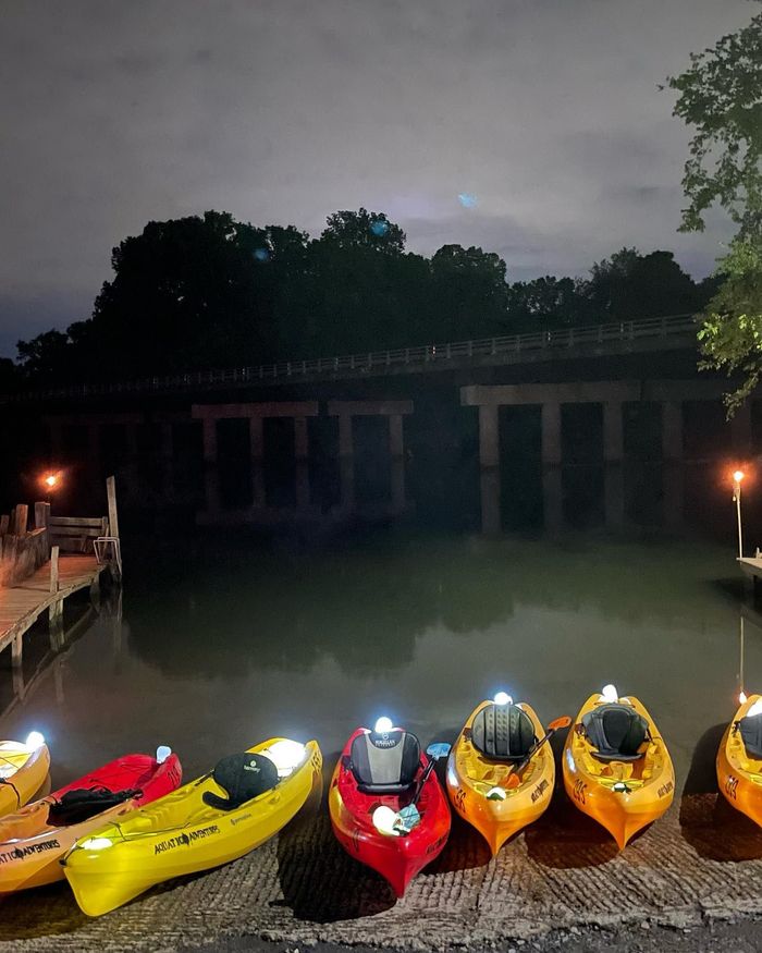 Kayaks lined up at a dock under a bridge at night; yellow and red boats with lights reflect on the dark water.