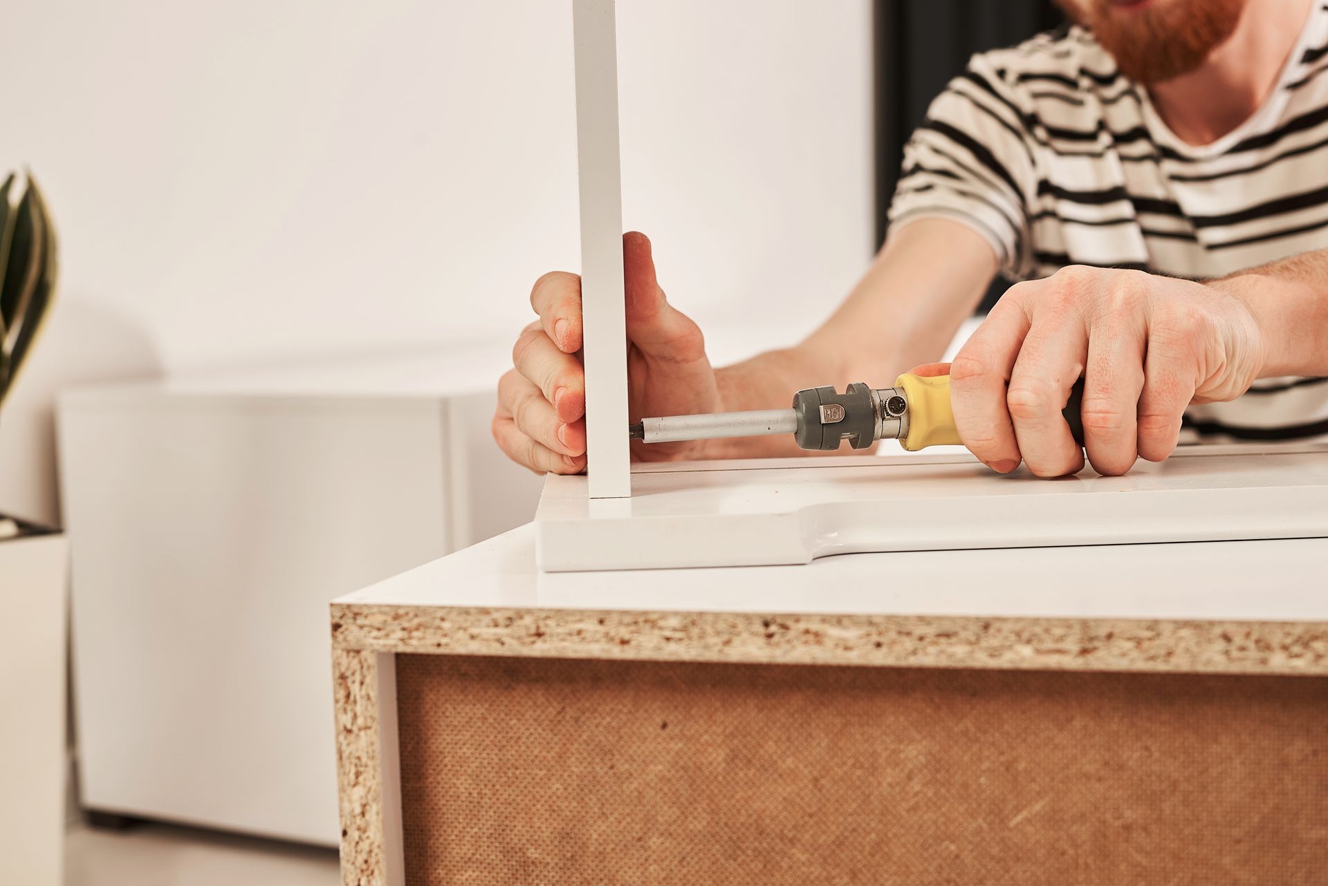 A person in a striped shirt uses a screwdriver to assemble pieces of white furniture on a wooden surface.