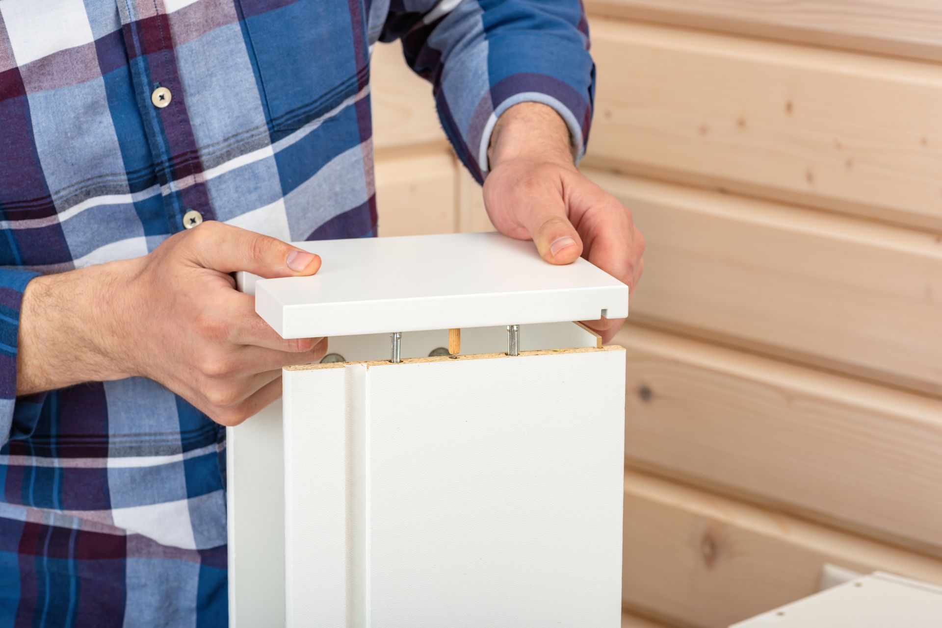 A person in a blue plaid shirt assembles white furniture panels, aligning a top piece with metal connectors.