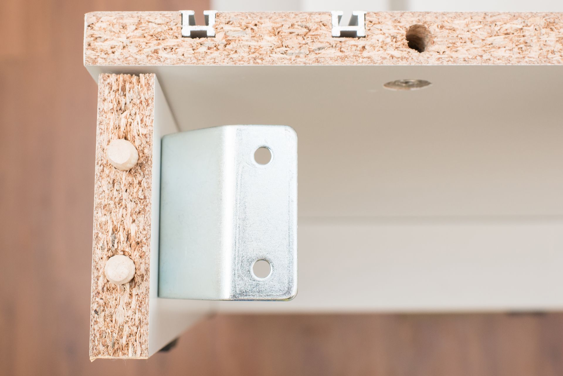 A metal corner bracket is installed on particle board furniture panels joined with two white dowels.