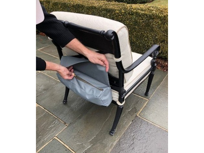 A person unzips a grey zippered bag attached to the back of a black and cream outdoor chair on a stone patio.
