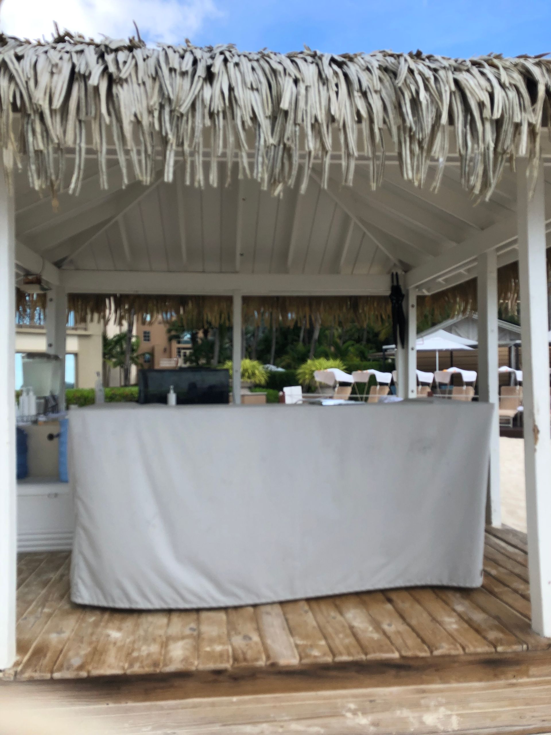 Beach bar with a white covered counter under a thatched roof, on a wooden deck, with ocean view in the background.
