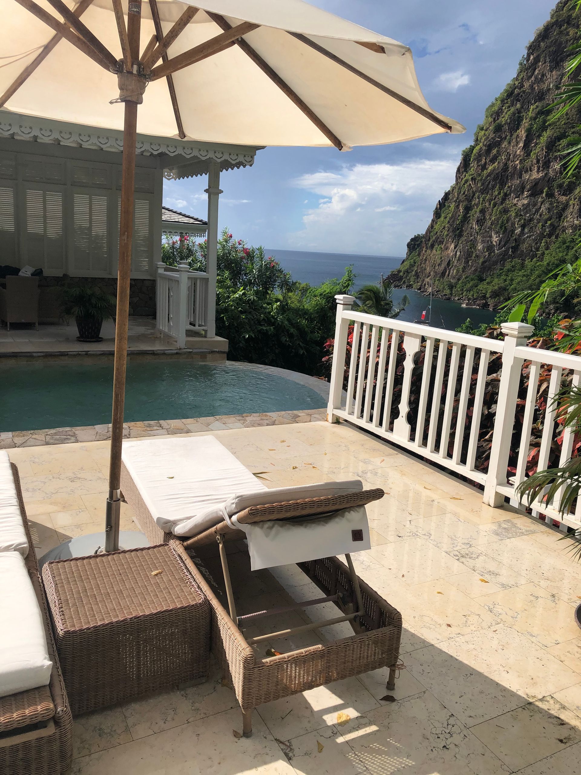A resort pool with an ocean view. A white umbrella shades a wicker lounge chair. A cliffside is visible in the background.