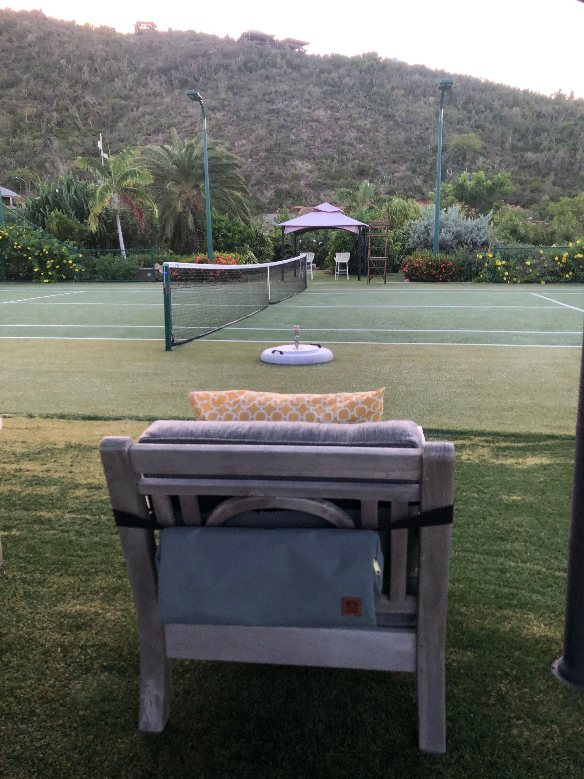 Wooden chair on a green lawn overlooking a tennis court with a mountain backdrop.