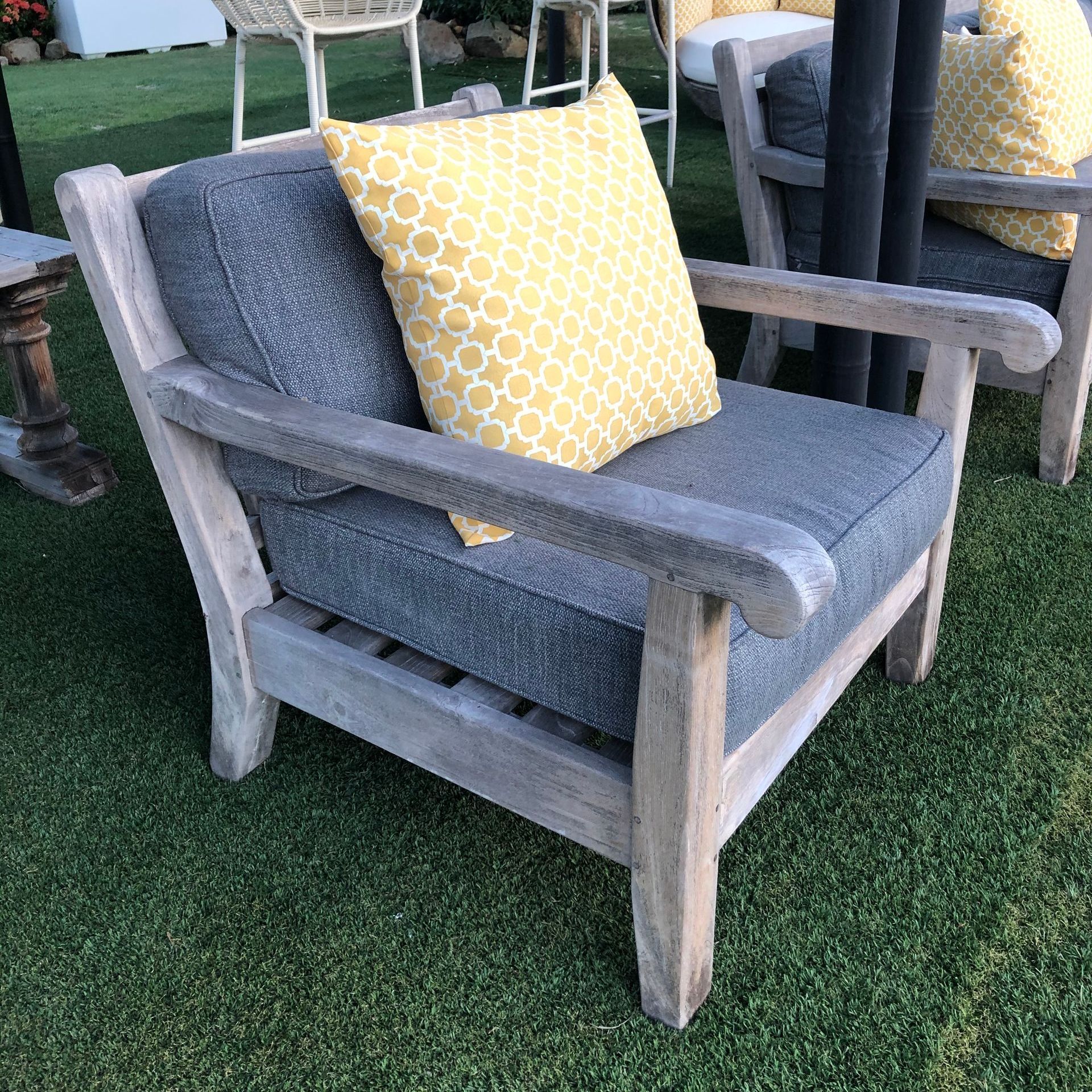 A weathered wood armchair with blue cushions and a yellow patterned pillow sits on green grass.