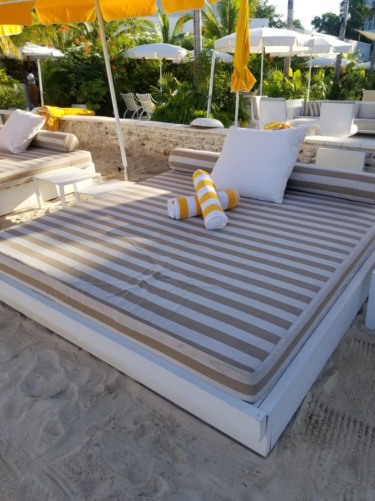 A beach daybed with striped beige and white cushions, white pillows, and yellow rolled pillows.