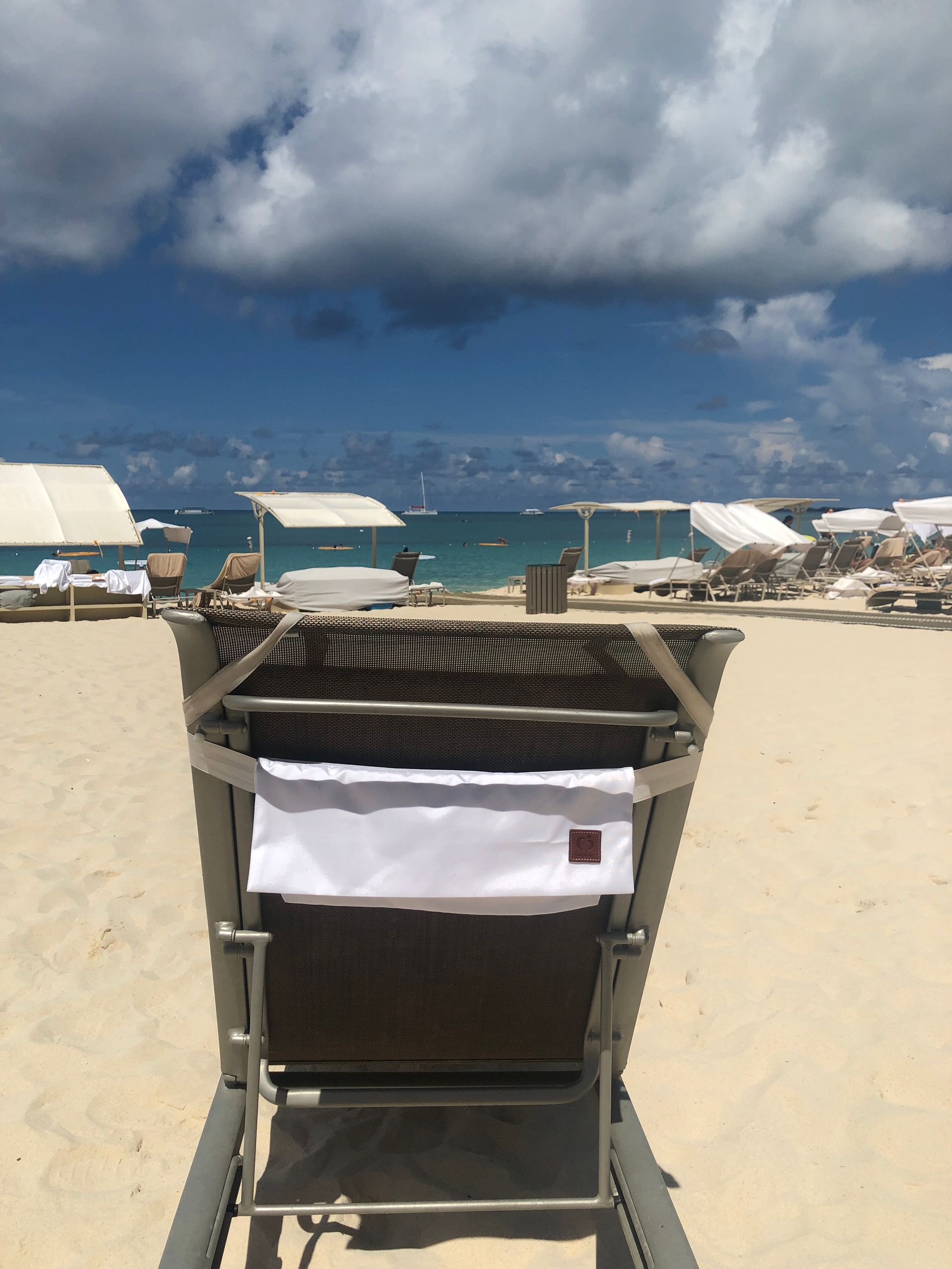 A beach scene with a wicker chair in the foreground, white sand, blue ocean, and a cloudy sky.