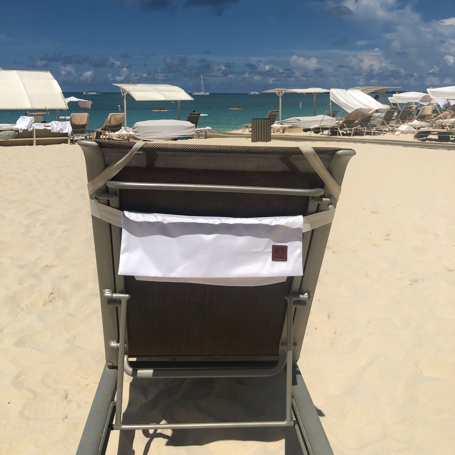 A beach chair on a sandy beach, facing the turquoise ocean. Other lounge chairs with canopies are in the background.