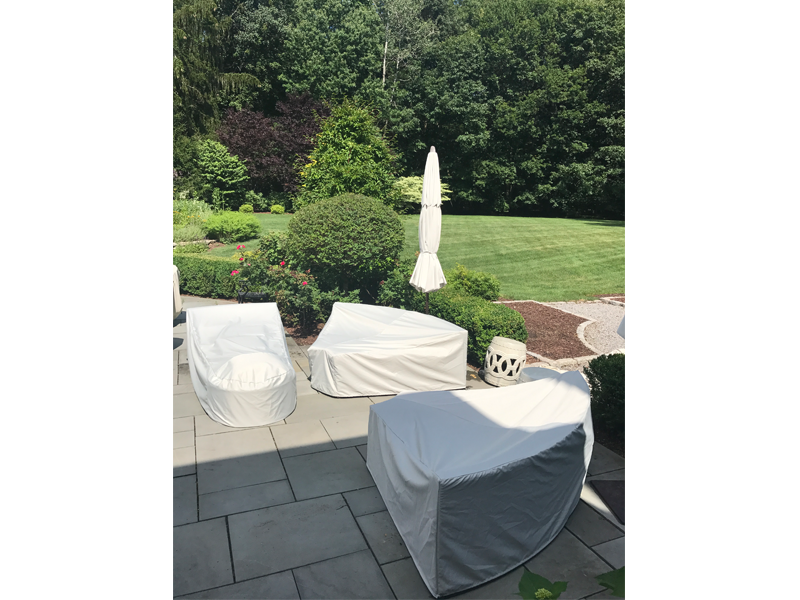 Patio furniture covered in light beige covers, with an umbrella, on a stone patio, overlooking a green lawn and trees.