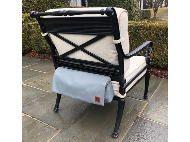 Back view of a black outdoor chair with light cushions, with a gray storage pouch attached. The chair is on a brick patio.
