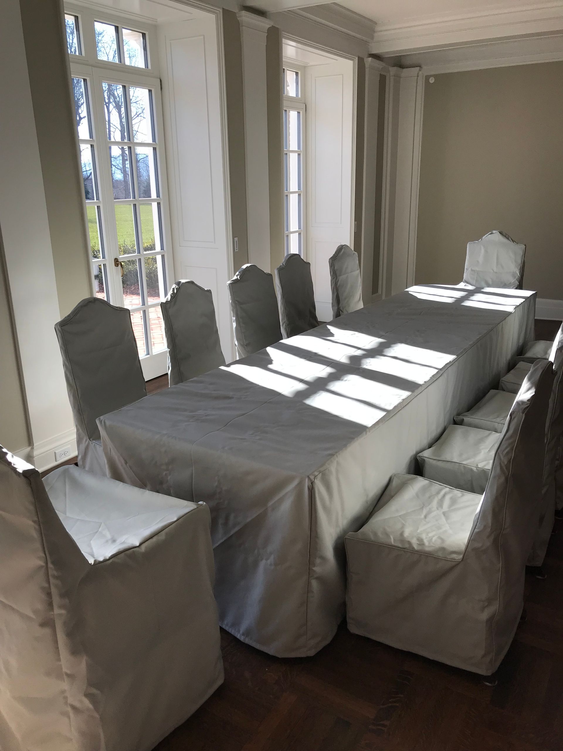 A dining room with a long white-covered table and matching chairs, set near large windows with striped curtains.