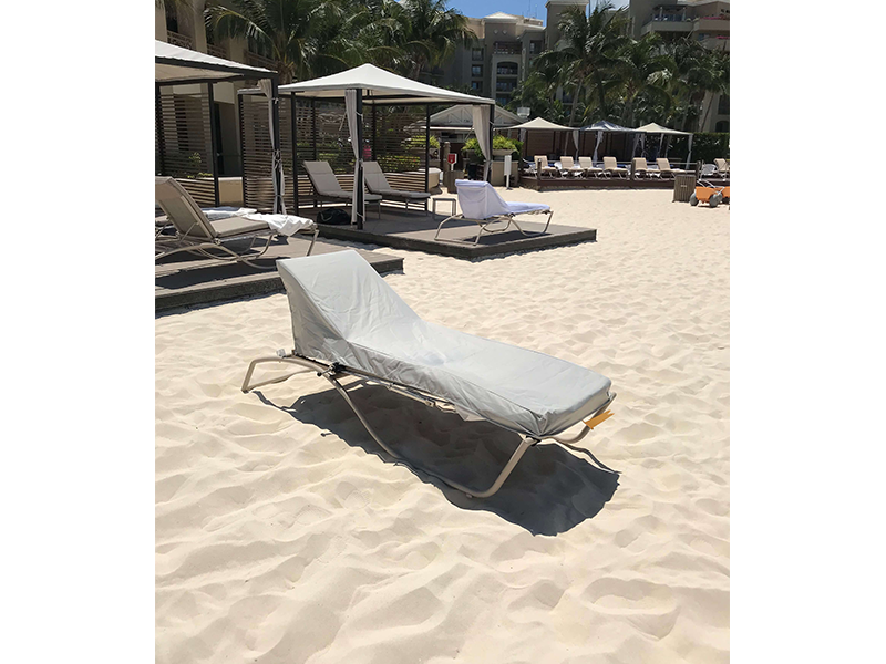 A grey-covered beach chair on a sandy beach, with cabanas and other lounge chairs in the background.