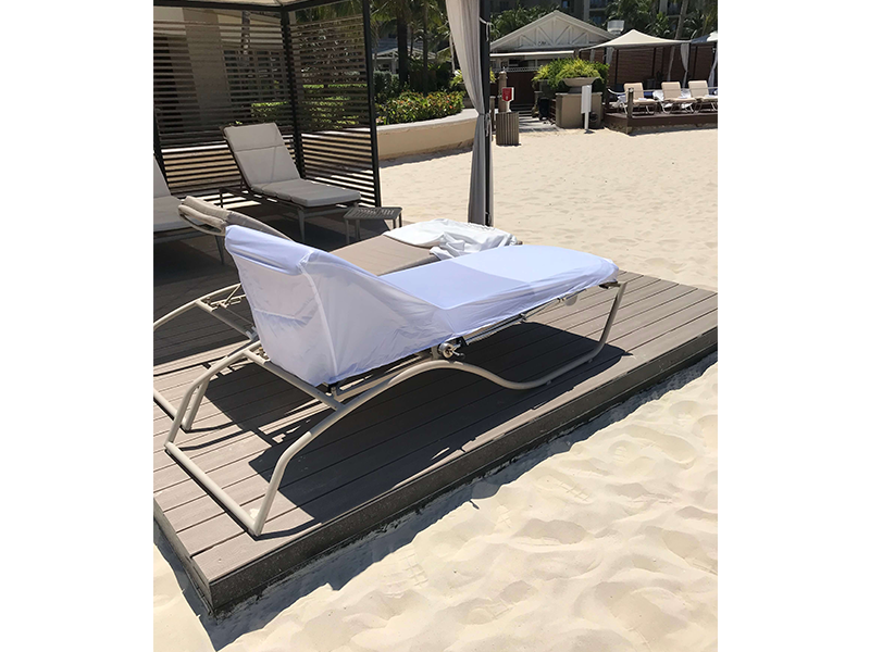 A lounge chair with a white cover sits on a wooden platform, near sand and other beach furniture.