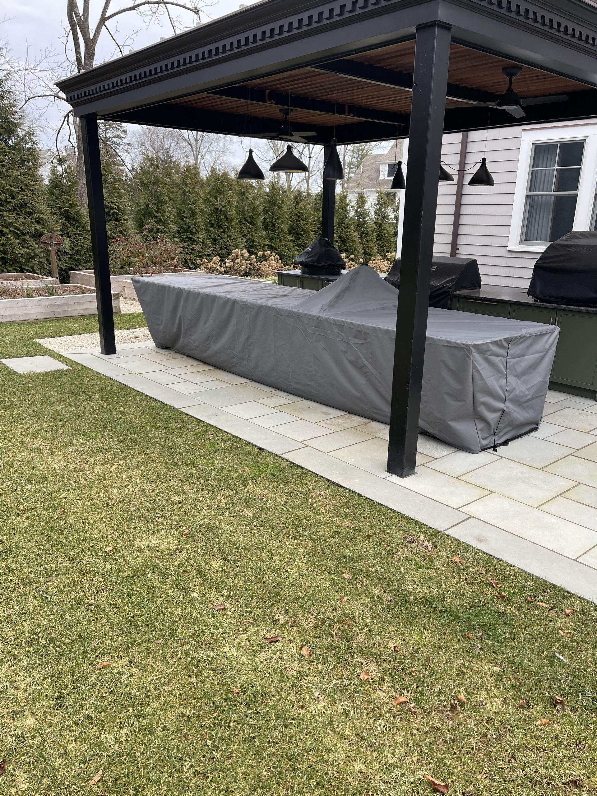 Outdoor kitchen setup with black pergola, gray countertop covered with tarp, and green grass.