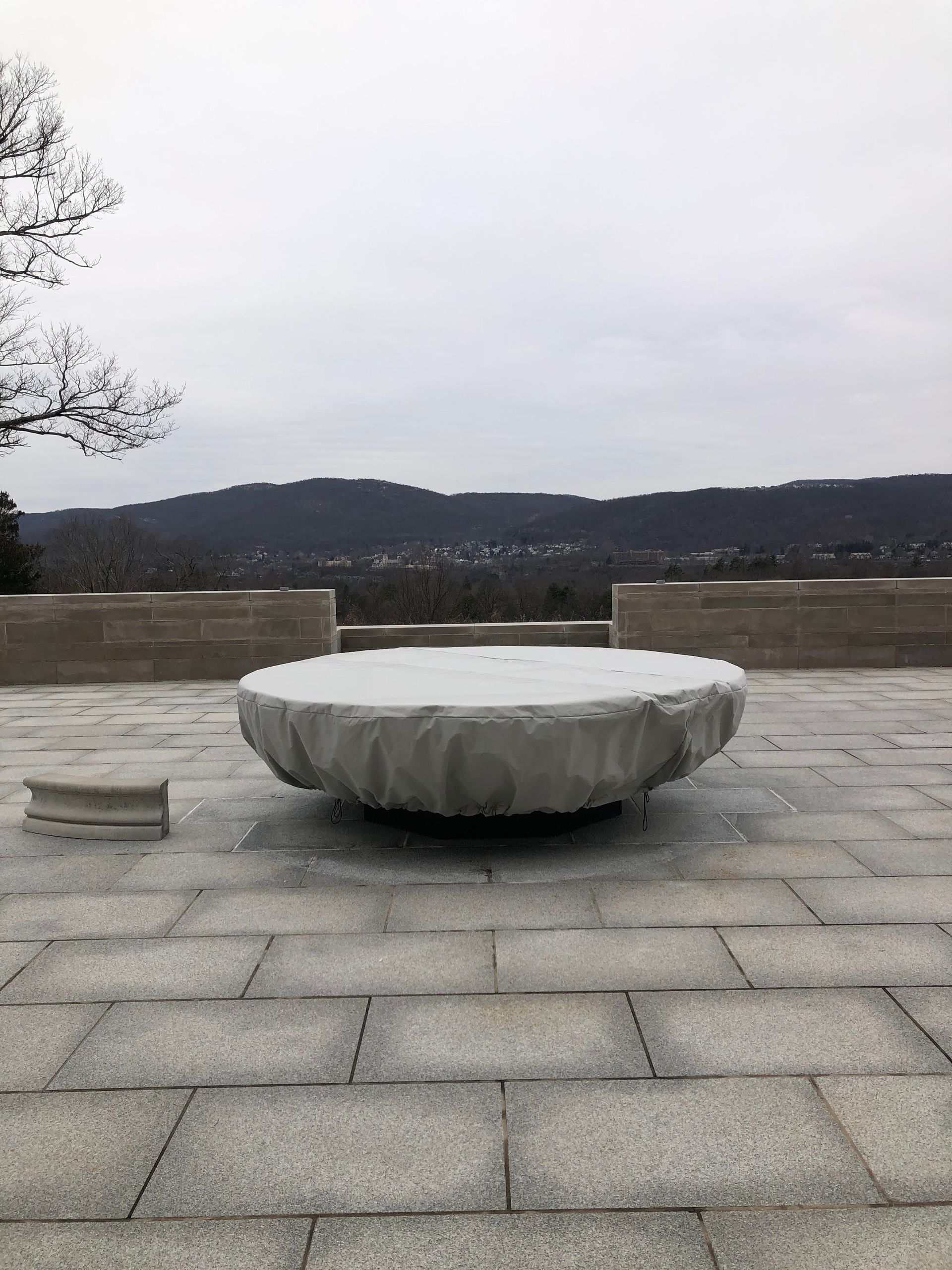 A large, covered circular structure on a stone patio, overlooking a distant tree-lined mountain range under a cloudy sky.