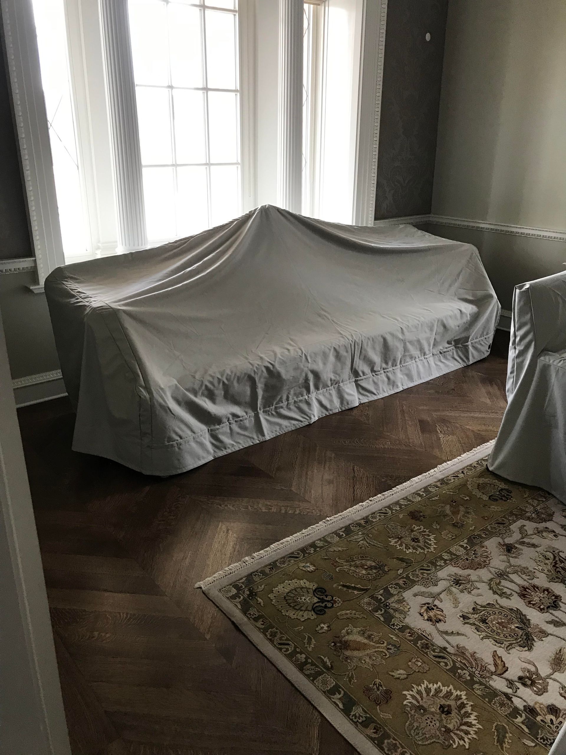 Living room with furniture covered in white sheets. A patterned rug sits on dark hardwood floors near a window.