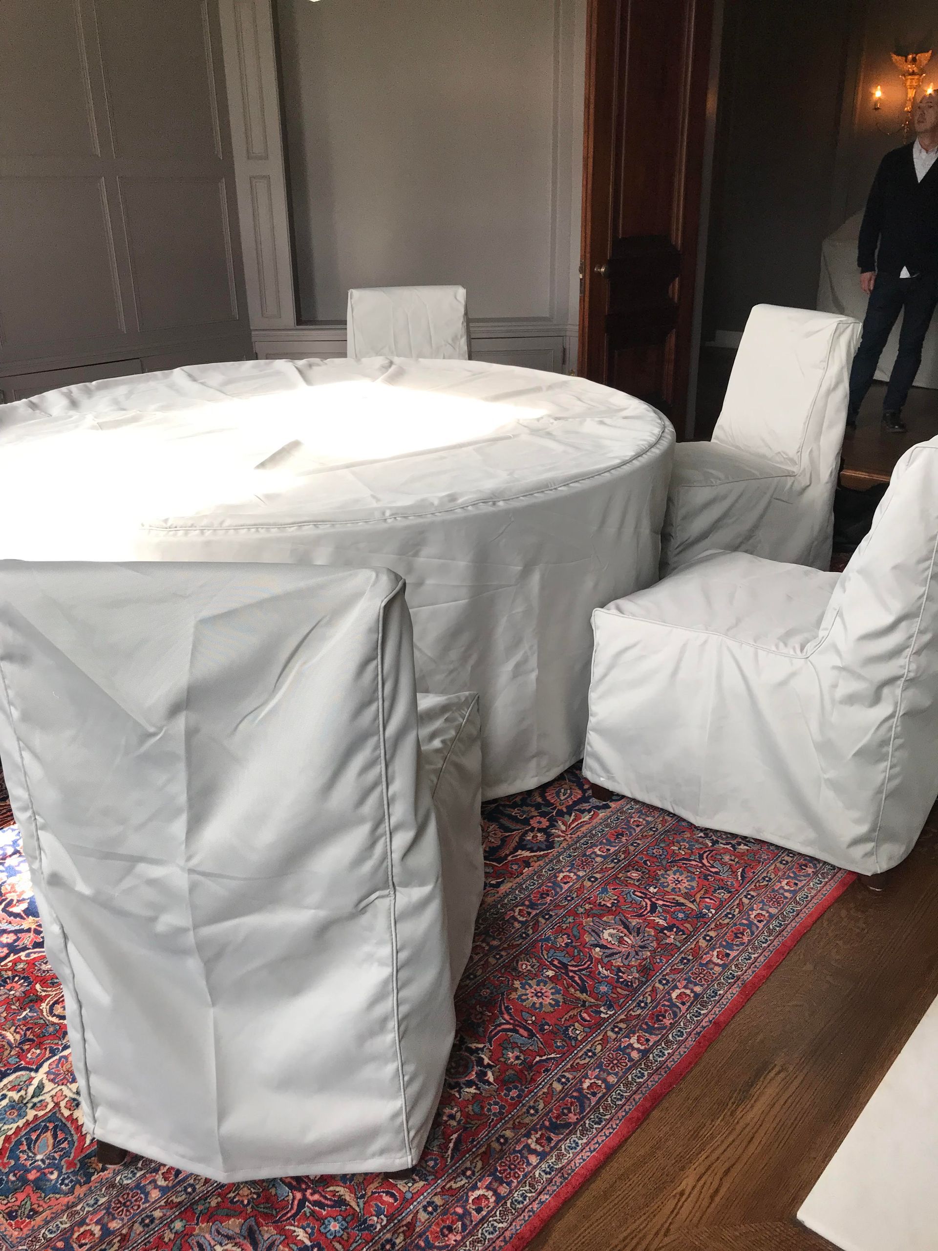 Round table and chairs draped in white covers, set on a patterned red rug. A person stands near a doorway.