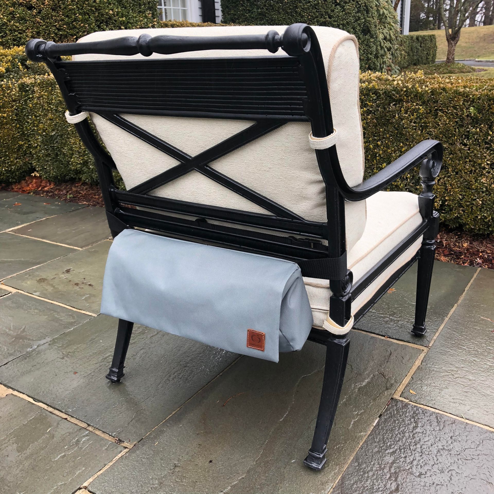 Back of an outdoor chair with a folded gray blanket attached. Chair is black with cream-colored cushions, on a stone patio.