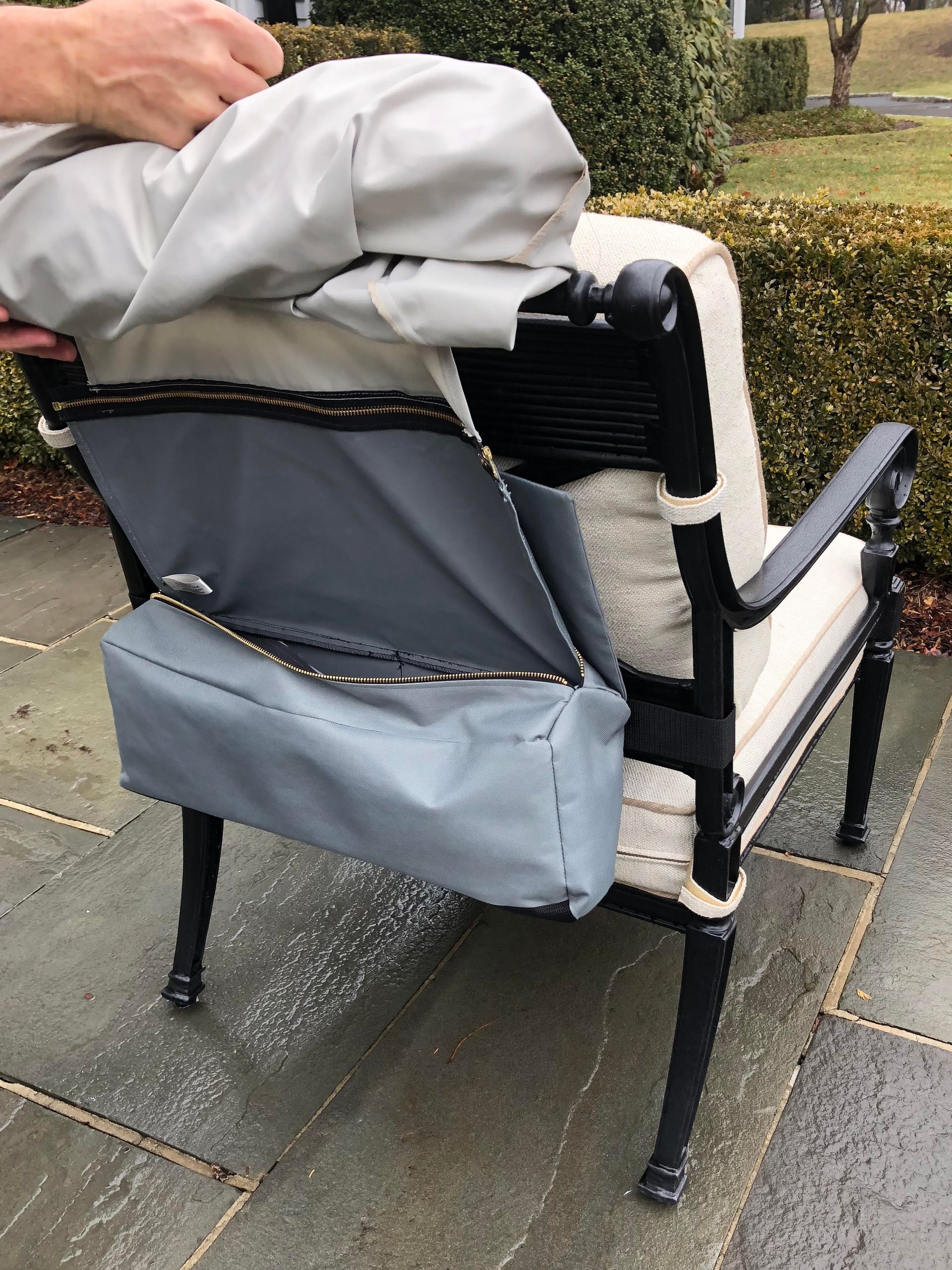 Person putting a gray cover over an outdoor chair with a storage pocket on the back, on a brick patio.