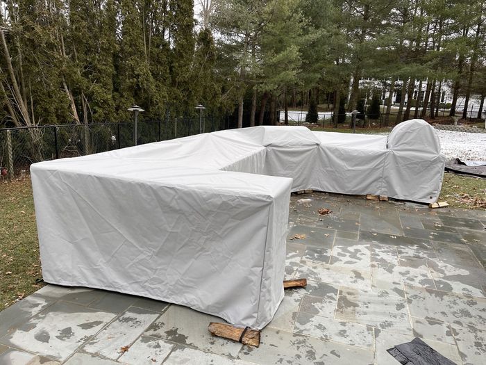 Covered outdoor furniture on a stone patio, surrounded by grass and trees; neutral-colored covers protect the seating.