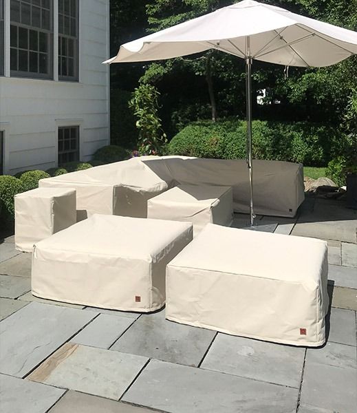 Outdoor patio furniture covered with light beige protective covers, under a white umbrella on a stone patio.