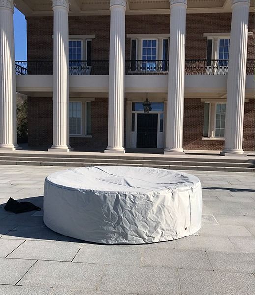A covered round hot tub sits on a stone patio in front of a two-story brick building with large white columns.