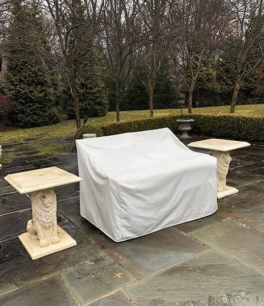 Outdoor furniture covered with a white tarp sits on a patio. Two square tables with lion base legs flank the covered couch.