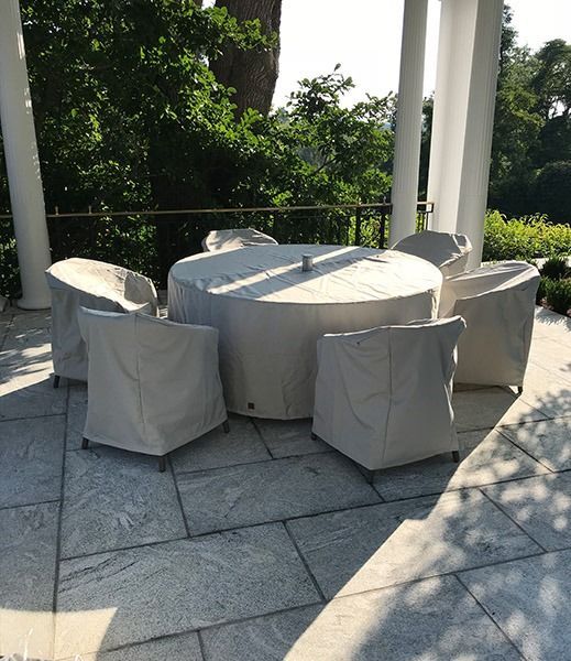 Patio furniture set covered in light beige protective covers on a gray stone patio, surrounded by white columns and greenery.