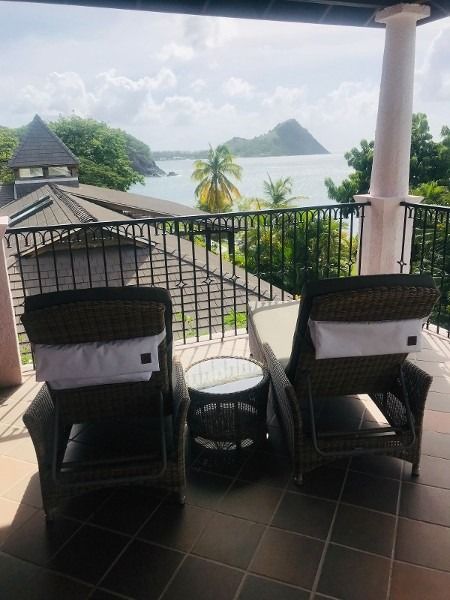 Two wicker chairs with white cushions sit on a balcony overlooking a bay with islands.