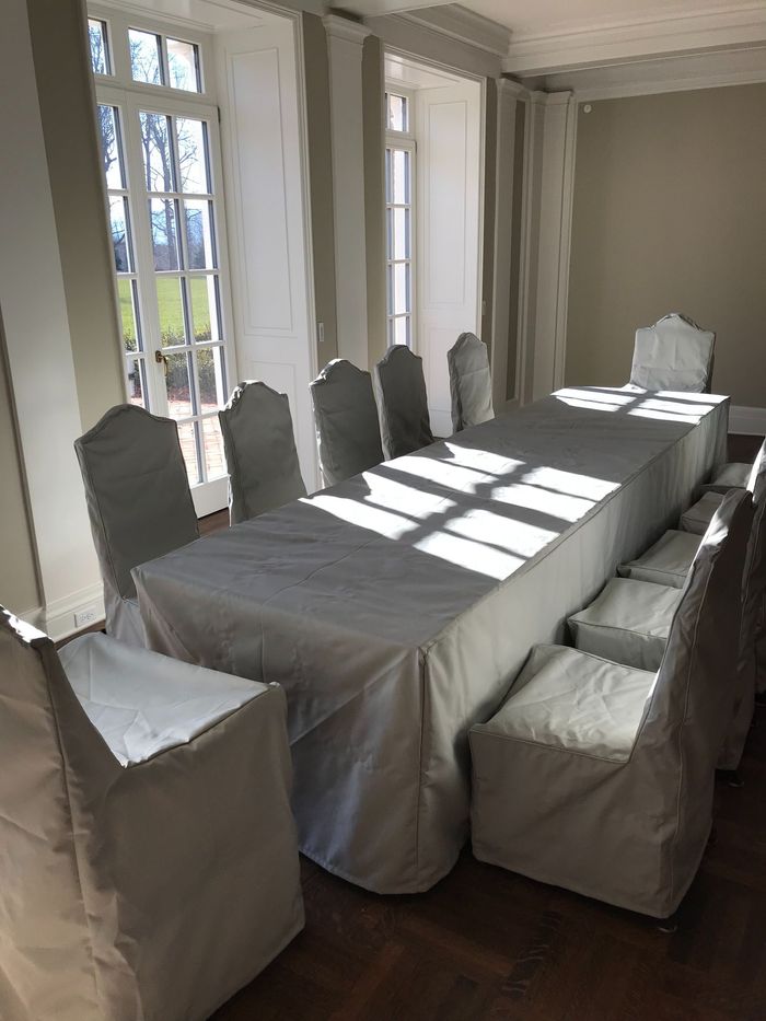 Dining room with a long white-covered table and matching chairs. Sunlight streams through the tall windows.